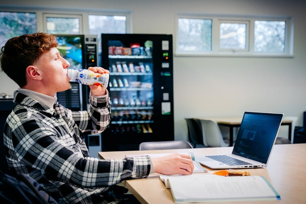Student drinkt een zuiveldrank uit de automaat terwijl hij aan zijn laptop werkt
